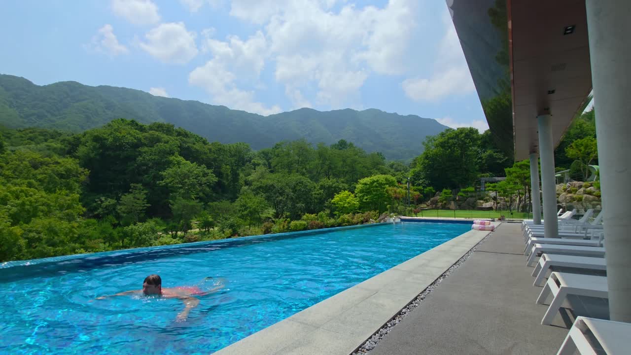 A fit man in his 30s enjoys swimming alone in a scenic infinity pool at a Gapyeong resort, surrounded by lush greenery and mountains, capturing the essence of summer relaxation
