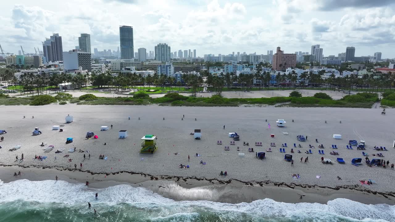 South Beach shoreline with beachgoers and umbrellas. Miami skyline in the distance with palm trees lining the coast. Aerial view of the bustling beachfront and nearby cityscape.