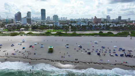 south beach shoreline with beachgoers and umbrellas. miami skyline in the distance with palm trees lining the coast. aerial view of the bustling beachfront and nearby cityscape.