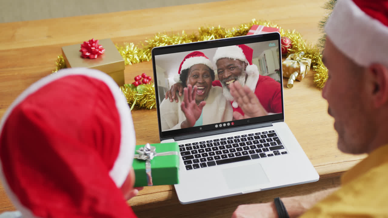 padre y hijo caucásicos con sombreros de santa usando una computadora portátil para una videollamada de navidad con una pareja en la pantalla