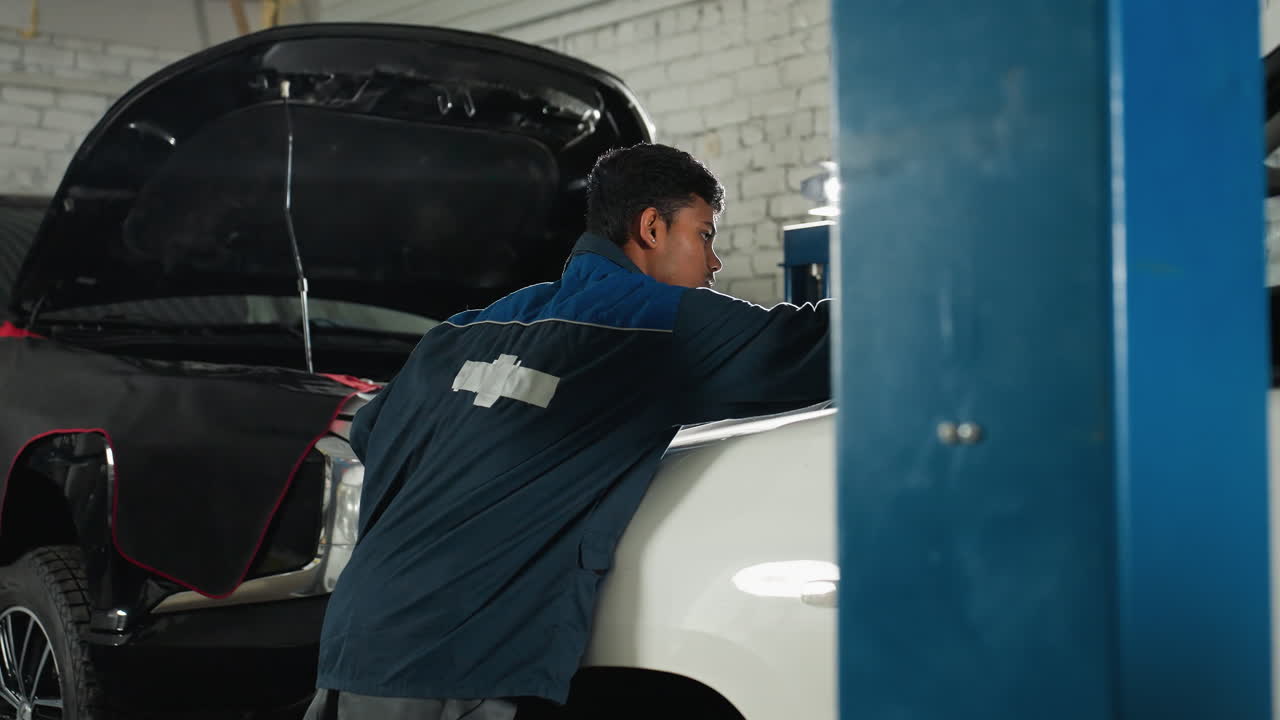 ingeniero de automóviles en uniforme azul arreglando algo en un coche blanco en un taller de automóviles, la luz brillando alrededor con un coche negro en el fondo, realizando reparaciones de vehículos en el garaje