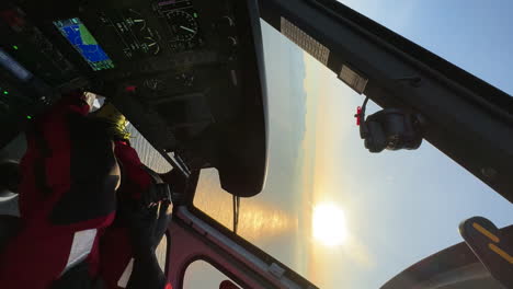 Vertical panning shot inside of a helicopter as it flies over a calm sea with the setting sun casting a golden light and snow covered islands in the distance