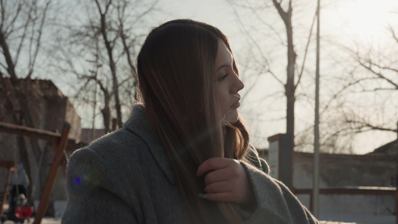 Bella donna che si spazzola i capelli all'aperto retroilluminata dalla luce solare, che indossa un cappotto di lana e tiene una spazzola rosa, seduta vicino a un'altalena del parco giochi, profilo tranquillo e contemplativo, atmosfera invernale fredda, morbida ora d'oro.
