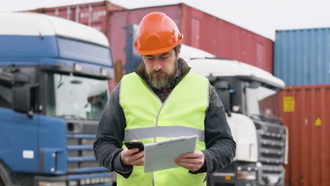 lavoratore che indossa giubbotto e casco di sicurezza che organizza una flotta di camion in un parco logistico mentre consulta un documento e parla al telefono 1