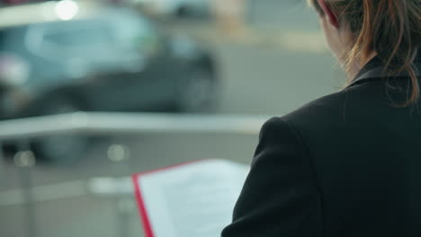 Close up rear view of woman in black suit reading file while standing beside metal railing with blurred view of parked cars and road