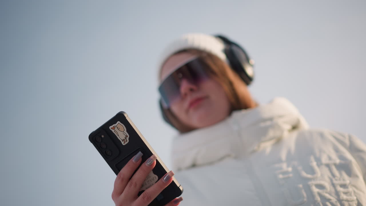 Fair skinned girl wearing tinted sunglasses and headphones twirling while holding smartphone in one hand scrolling screen in snowy urban park gentle sway to music beats stirring joy