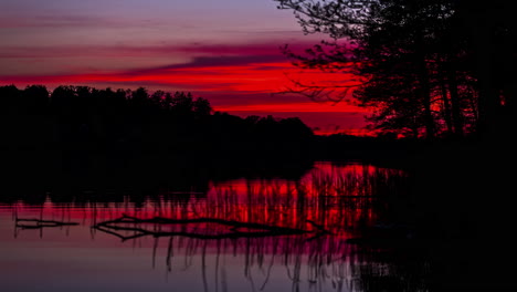 Static-view-of-red-sky-at-sunset-over-water-in-timelapse,-with-trees-on-the-bank-of-the-lake-in-the-evening-with-sun-setting-over-the-horizon