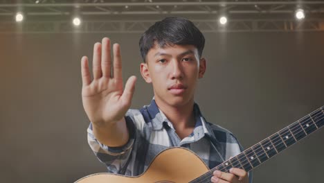 primer plano de un adolescente asiático con una guitarra haciendo un gesto de parada mientras está de pie en el escenario