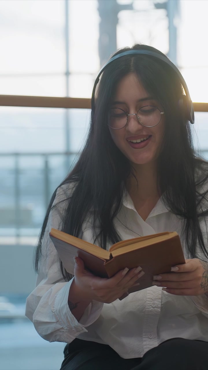 Mujer joven con cabello largo y oscuro lee un libro. La mujer sostiene un libro abierto de cubierta marrón y pasa las páginas mientras se concentra. La luz diurna brillante entra por los cristales de las ventanas, revelando un fondo borroso.