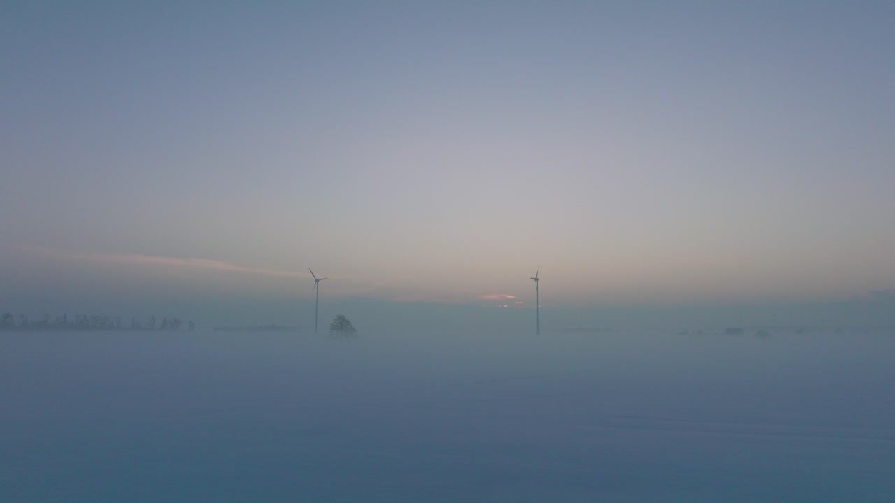 풍력 발전소에서 재생 에너지를 생성하는 풍력 터빈의 공중 전망, 안개로 가득 찬 눈으로 가득 찬 시골 풍경, 황금 시간의 빛이 있는 화창한 겨울 저녁, 넓은 드론 샷이 앞으로 낮게 움직입니다.