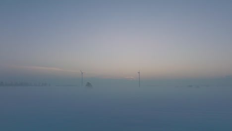 Aerial-view-of-wind-turbines-generating-renewable-energy-in-the-wind-farm,-snow-filled-countryside-landscape-with-fog,-sunny-winter-evening-with-golden-hour-light,-wide-drone-shot-moving-forward-low