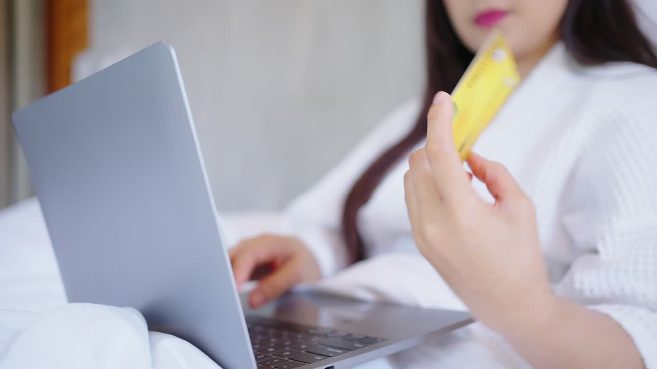 아시아 여성이 노트북 컴퓨터와 은행 신용카드로 온라인 쇼핑을 하고, 침실의 편안함 속에 있는 여성