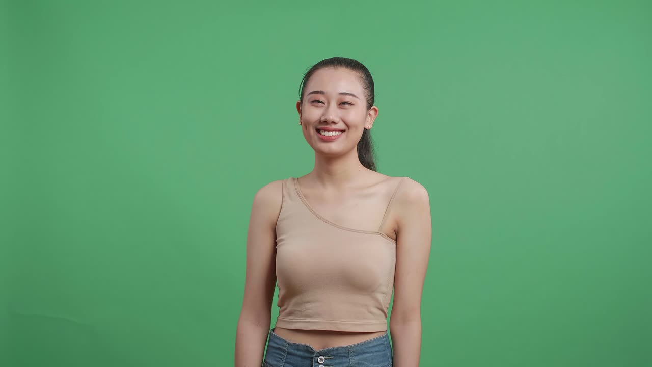 아시아 여성이 스튜디오에서 녹색 스크린 배경에 서서 카메라에 미소 짓고 있습니다.