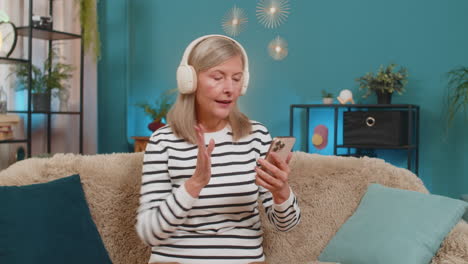 mature woman listening to music on smartphone with wireless headphones dancing resting on home sofa