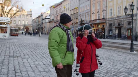 Senior-couple-tourists-grandmother-grandfather-taking-photo-pictures-on-retro-camera-in-winter-city