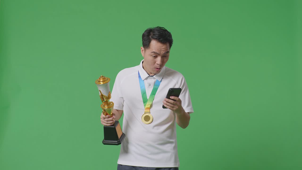 금메달과 트로피를 가진 아시아인 남자가 스마트폰을 보고 스튜디오의 녹색 화면 배경에서 와우라고 말합니다.
