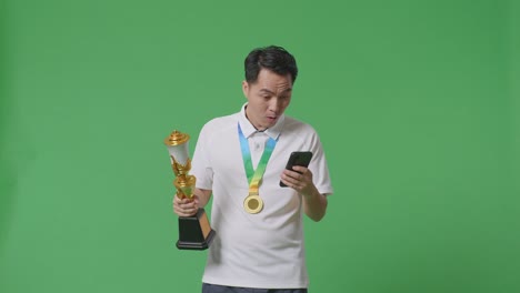 hombre asiático con una medalla de oro y un trofeo mirando su teléfono inteligente y luego diciendo wow en el fondo de la pantalla verde en el estudio