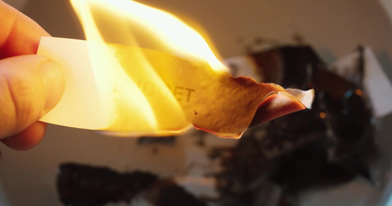 Et nærbillede i slow-motion af et stykke papir mærket “BUDGET”, der brænder. Flammen fortærer papiret gradvist og symboliserer økonomisk tab, overforbrug eller spild