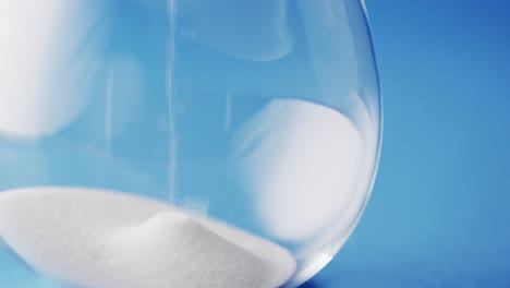 video of close up of hourglass with white sand pouring with copy space on blue background