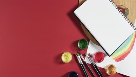 overhead view of notebook, paints and brushes on red background with copy space, in slow motion