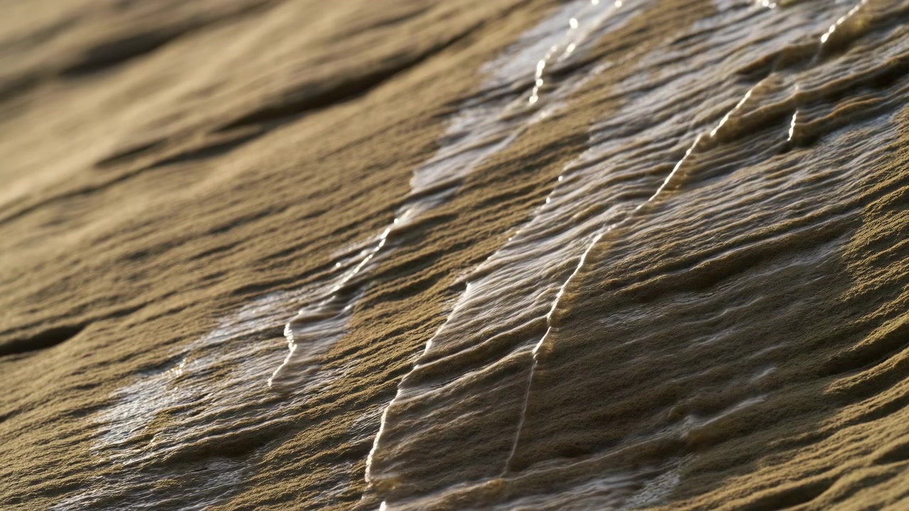 Vista diagonale ravvicinata di sabbia della spiaggia increspata con sottili veli traslucidi di acqua marina depositati lungo le creste e i canali, creando striature lucide, chiazze bagnate, tenui riflessi e una texture costiera naturale