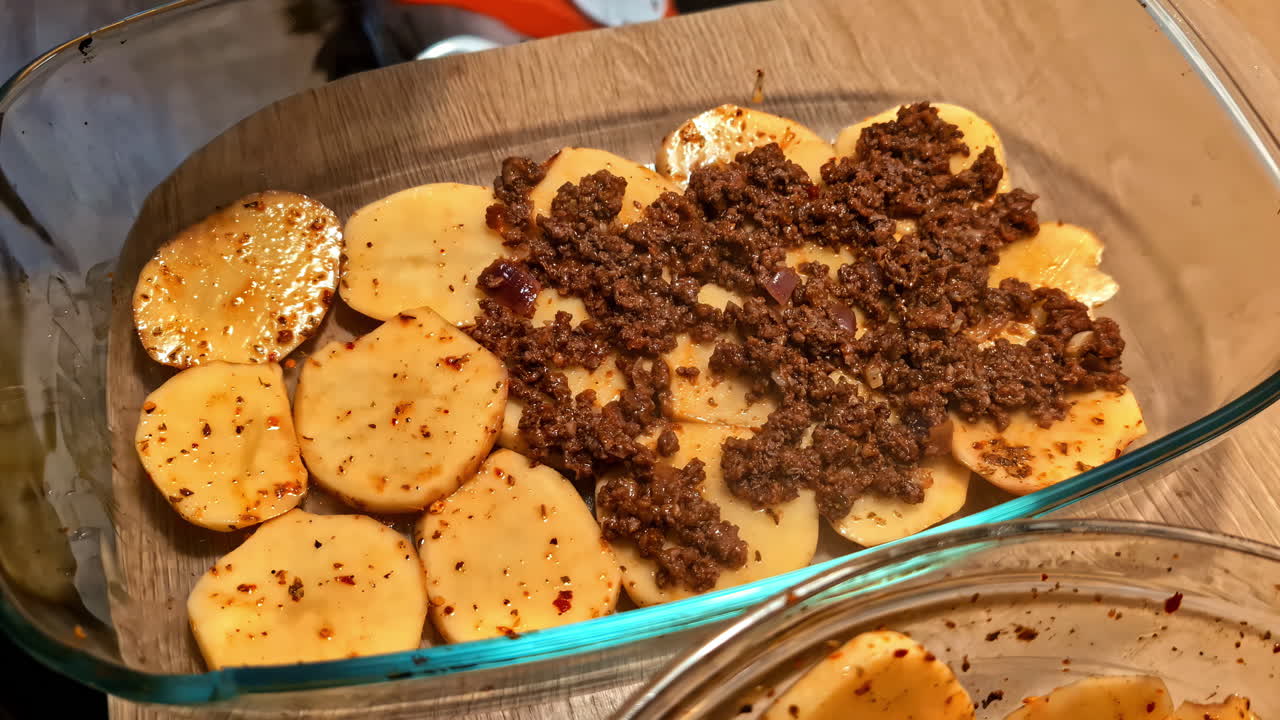 A close-up of hands layering seasoned ground beef over sliced potatoes in a glass baking dish, preparing a homemade casserole in a cozy kitchen in Latvia