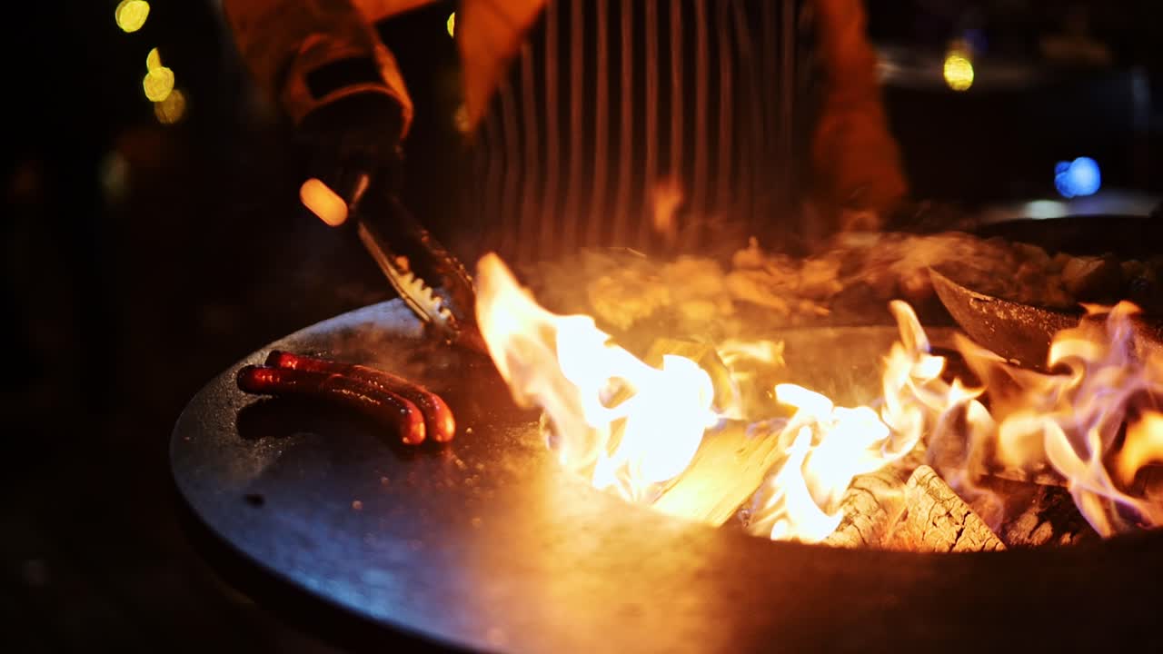 Holiday market chef prepares sausages over flames using tongs, rural Latvia
