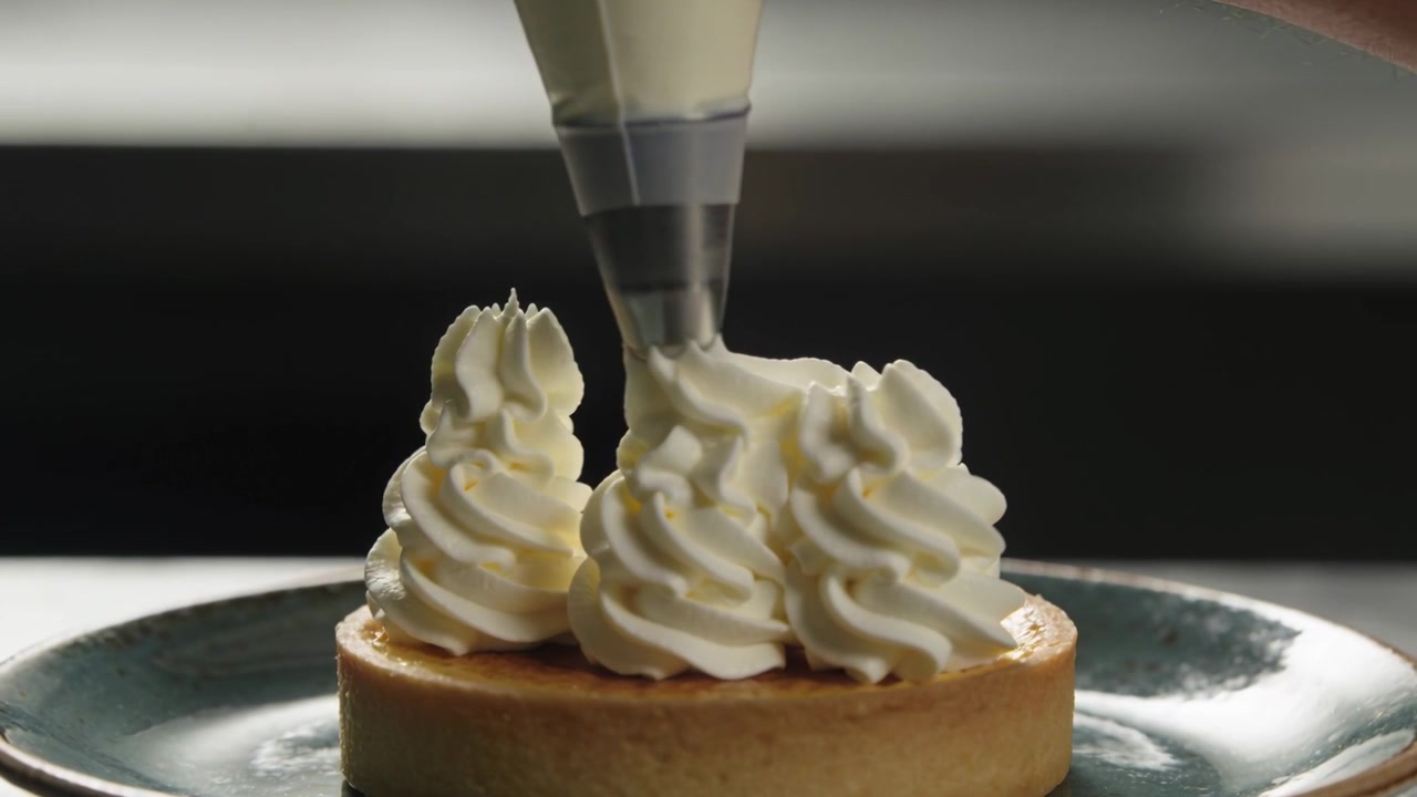 Squeezing piping bag, chef piping three cream swirls on tart at counter, guiding nozzle, decorating