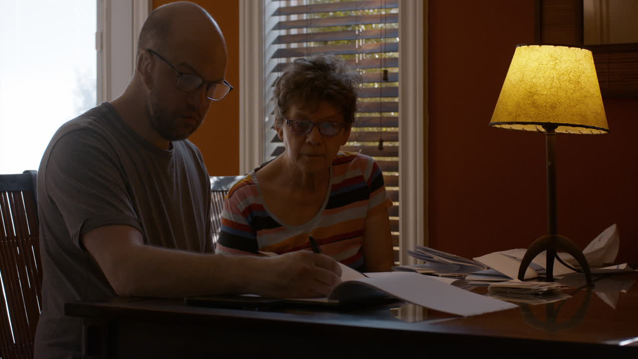 An adult son and his elderly mother sit together at a dining table in warmly lit home, sorting through papers and bills. The son jots down notes in a notepad, sifting papers. Onlooking mother directs.