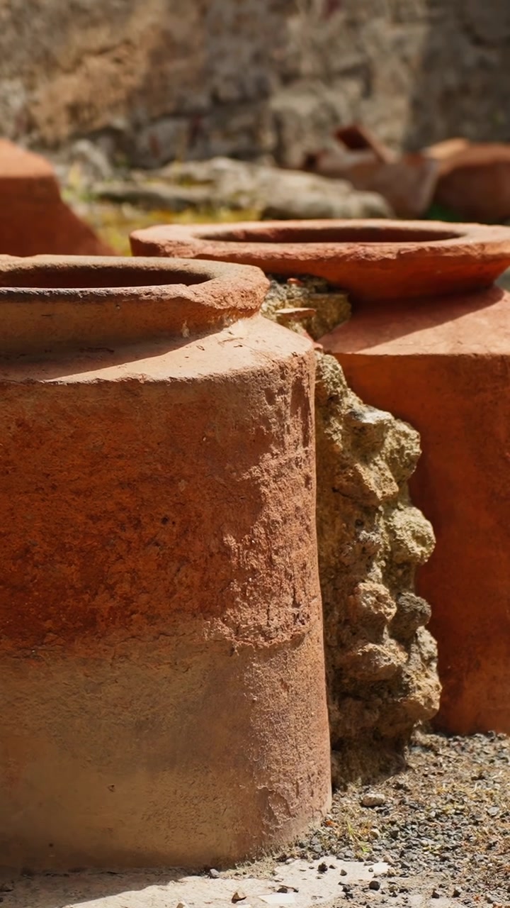 Vertical close-up of terracotta storage jars in Pompeii ruins, with rough clay texture, gravel floor, sunlight, and ancient Roman courtyard pottery details.