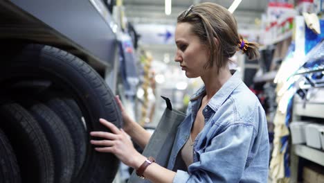 Linda-Chica-En-Un-Gran-Centro-Comercial-Seleccionando-Neumáticos-De-Coche.-Examen.-Comprador.-Filas-De-Diferentes-Neumáticos-De-Coche.-Vista-Lateral