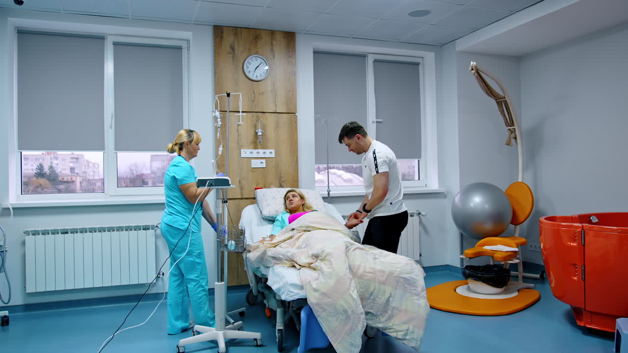 Pregnant lady lies in hospital bed. Nurse attaches drop counter to woman's hand. Husband stands beside soothing his wife.