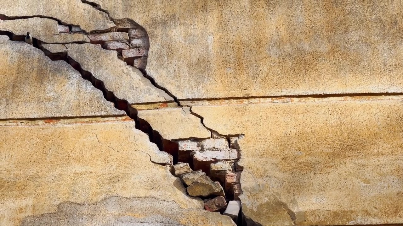 Severe crack in an old plaster wall with exposed brickwork, broken facade layers, weather stains, and visible structural damage in bright sunlight.