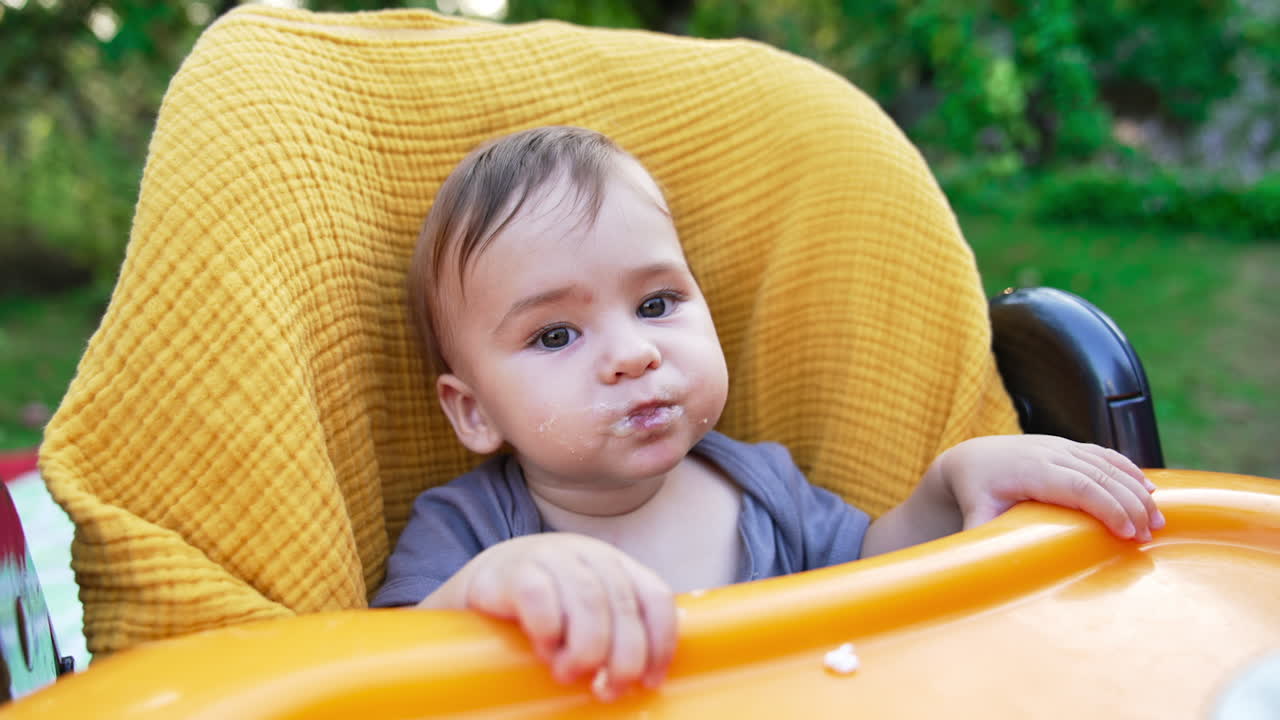Sweet lovely baby boy nutrition outdoors in summer. Caring mother feeds her son with porridge from a spoon. Blurred nature backdrop.