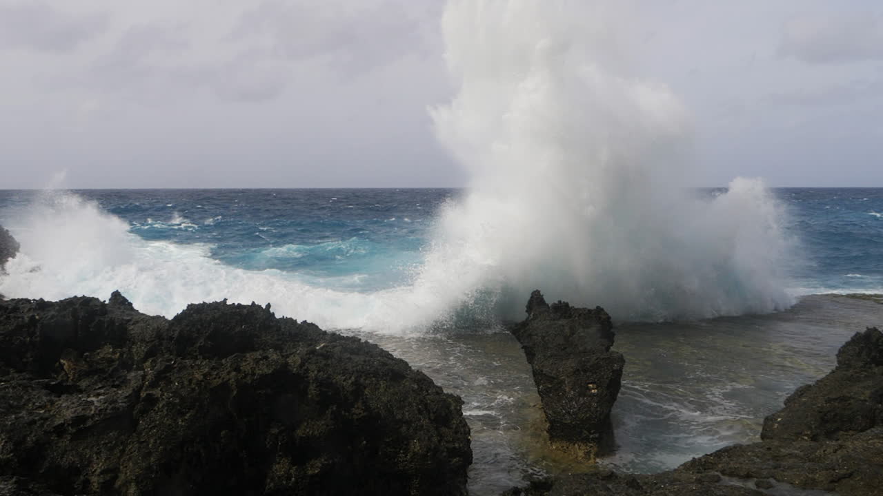 una ola oceánica choca contra las rocas a lo largo de la costa salpicando agua en el cielo y en la lente de la cámara - acción intensa en cámara lenta