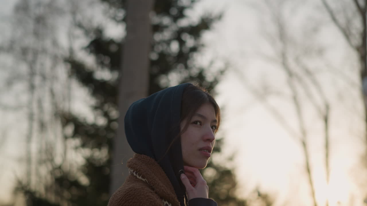 일몰에 공원을 걸어가는 슬픈 소녀의 클로즈업, 갈색 코트와 후드와 함께 후디를 입고, 그녀의 얼굴은 부분적으로 숨겨져 있으며, 부드럽고 희미한 빛에 대해 우울한 분위기를 반영합니다.