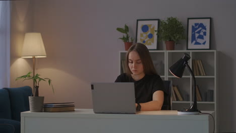 trabajadora remota mujer en su oficina de casa por la noche sentada en la mesa en la sala de estar trabajando con la computadora portátil hablando en el trabajo hablando escribiendo texto en el teclado