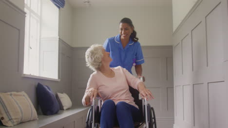 portrait of senior woman at home being pushed in wheelchair by female care worker in uniform