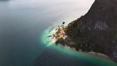 drone orbit over pinagbuyutan island, palawan, where steep limestone cliffs meet a narrow stretch of white sand lined with cocos nucifera palms and shallow and transparent waters teeming marine life