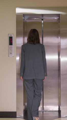 businesswoman in gray suit walking toward elevator with closed metallic doors on floor 3, viewed from behind in office hallway. scene reflects professional routine, workplace environment
