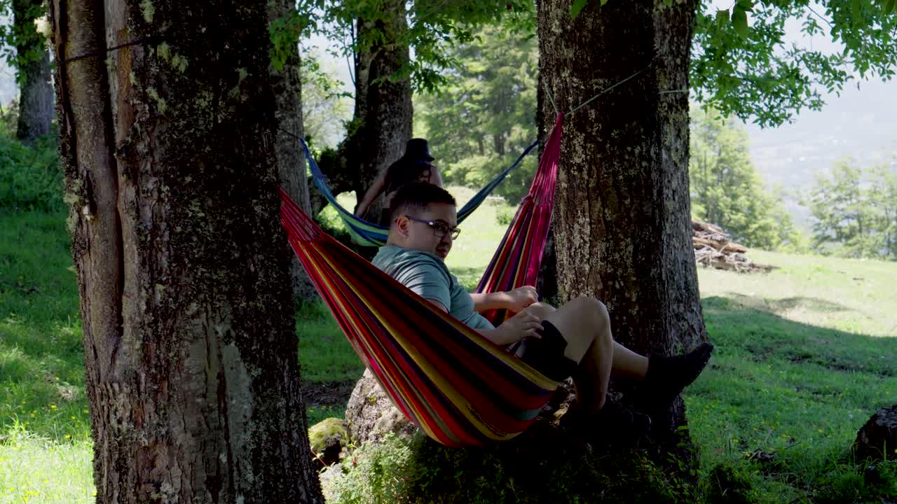 Handheld of man and woman resting in hammocks hanging from trees in green valley near Laguna Corazon in Liquiñe, Chile