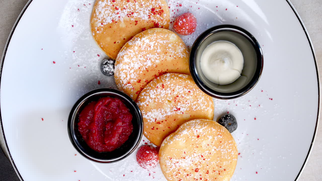 Small pancakes are served on a white plate with fresh raspberries and blueberries, sour cream and jam, a glass of orange juice is placed next to the plate on a gray background