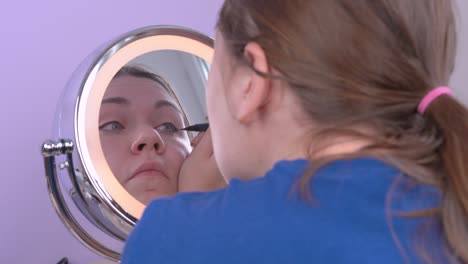 joven mujer atractiva aplica el delineador de ojos dibujando ojos de gato maquillaje, mirando su reflejo en el espejo redondo y tratando de hacerlo con precisión. disparar desde la espalda. fondo lila, en interiores.