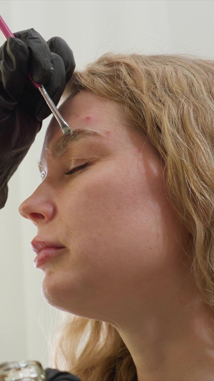 Close up view shows hand wearing black gloves applying makeup to eyebrow with pink brush. scene is set in bright clean environment that suggests careful and precise beauty treatment