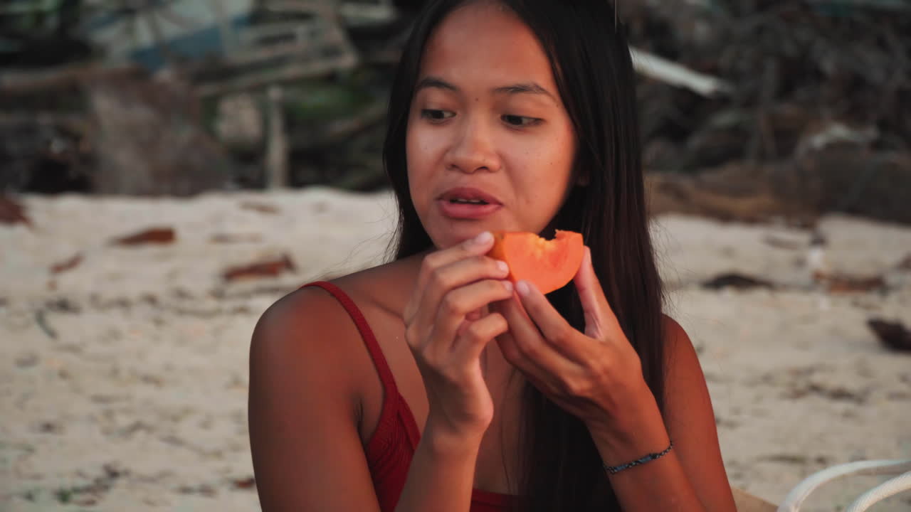 Giovane donna sulla spiaggia che mangia papaya, bagliore del tramonto, occhiali da sole appoggiati sulla testa, litorale sabbioso e sfondo di legni spiaggiati, morsi lenti e contemplativi, sorriso naturale, luce calda tropicale, degustazione intima.