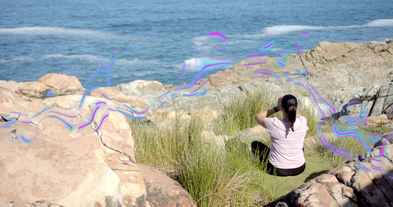 Donna di mezza età che alza le braccia iniziando una sequenza yoga su una costa rocciosa, guidando il respiro con le onde dell'oceano.
