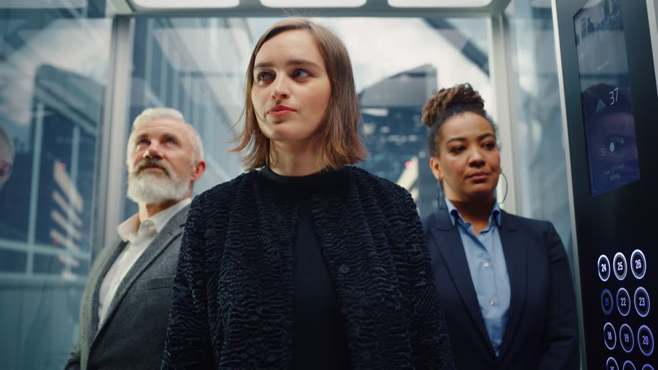 세 명의 다양한 다민족이 현대적인 비즈니스 센터에 있는 사무실로 유리 엘리베이터를 타고 올라가는데, 엘리베터 앞에 서 있는 행복한 젊고 아름다운 사업가에 초점을 맞추고 있다.