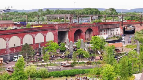 ascending aerial view of mayfield park in manchester, england, showing modern playground structures, lush greenery, and historic red-brick railway arches with passing trains and industrial details