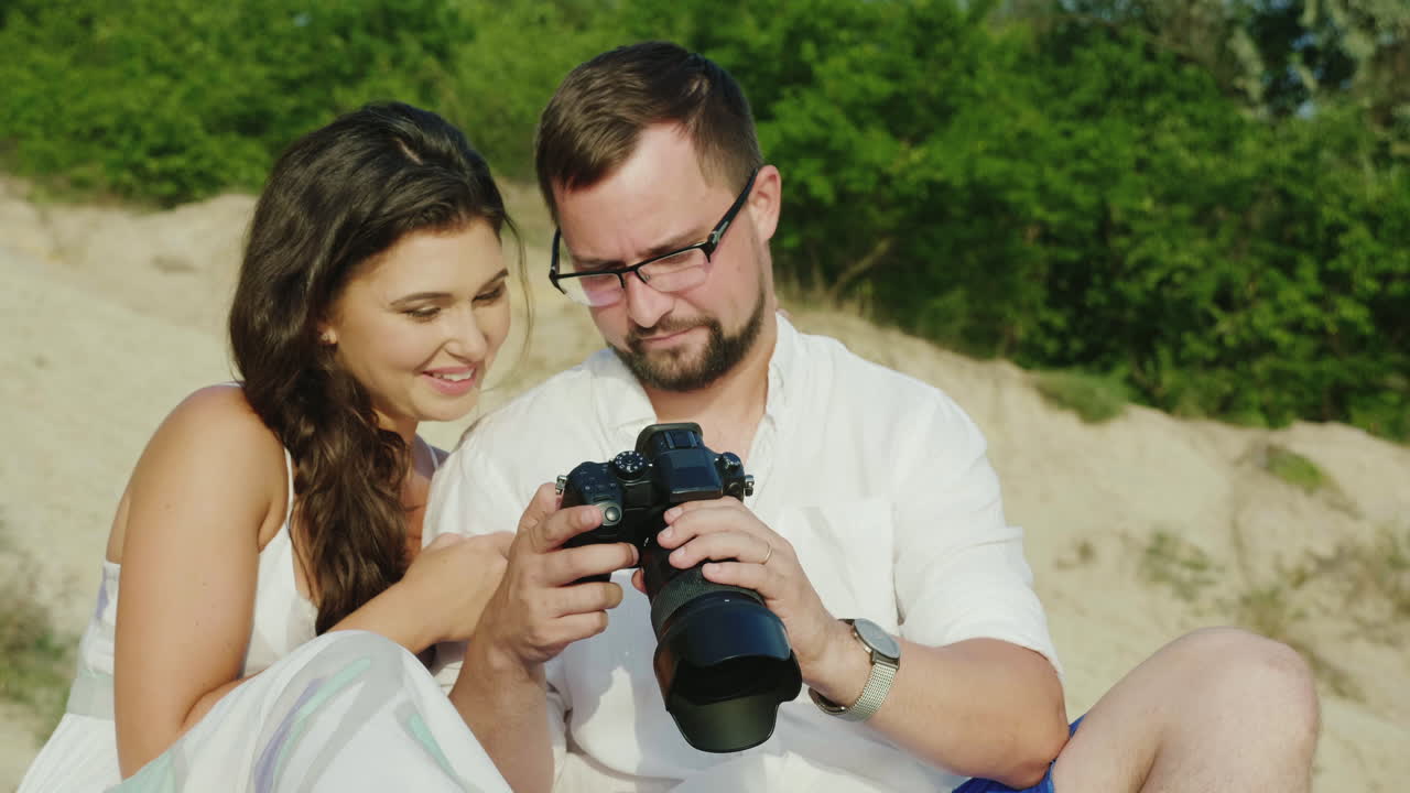 Um jovem casal olha as fotos em uma câmera digital, sentados perto da praia. Grandes lembranças.