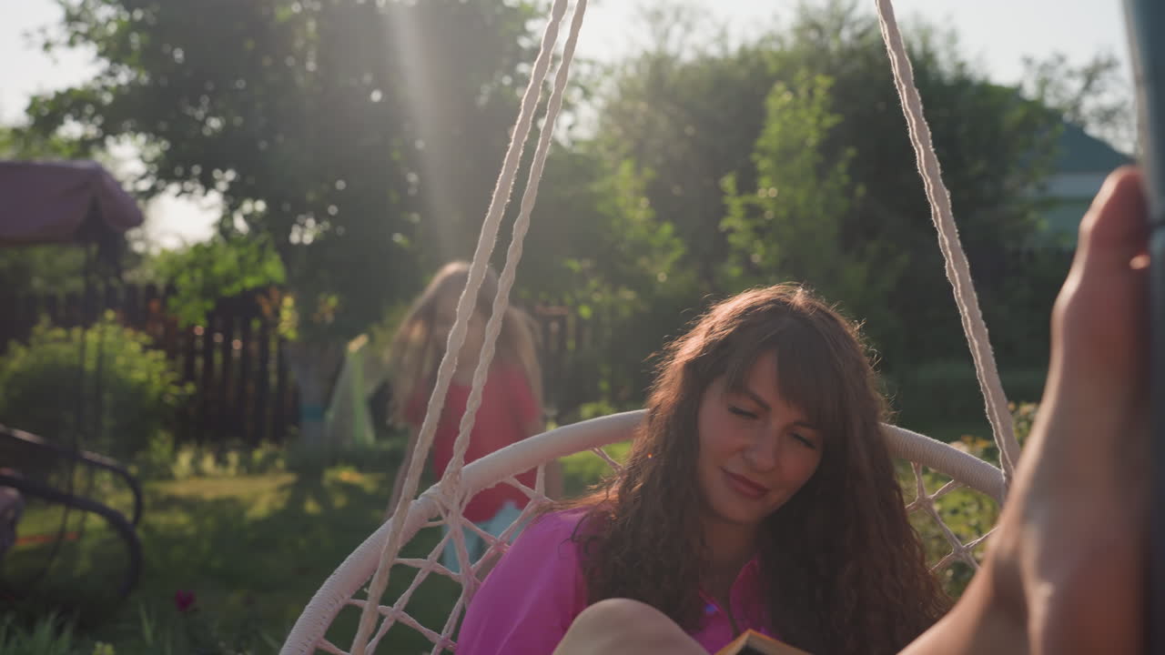 Sunlit Yard Captures Touching Family Interaction, Woman Relaxes On Swing As Child Gives Affectionate Kiss, Sunshine Bathes Backyard Where Woman Reads And Child Offers Loving Cheek Kiss